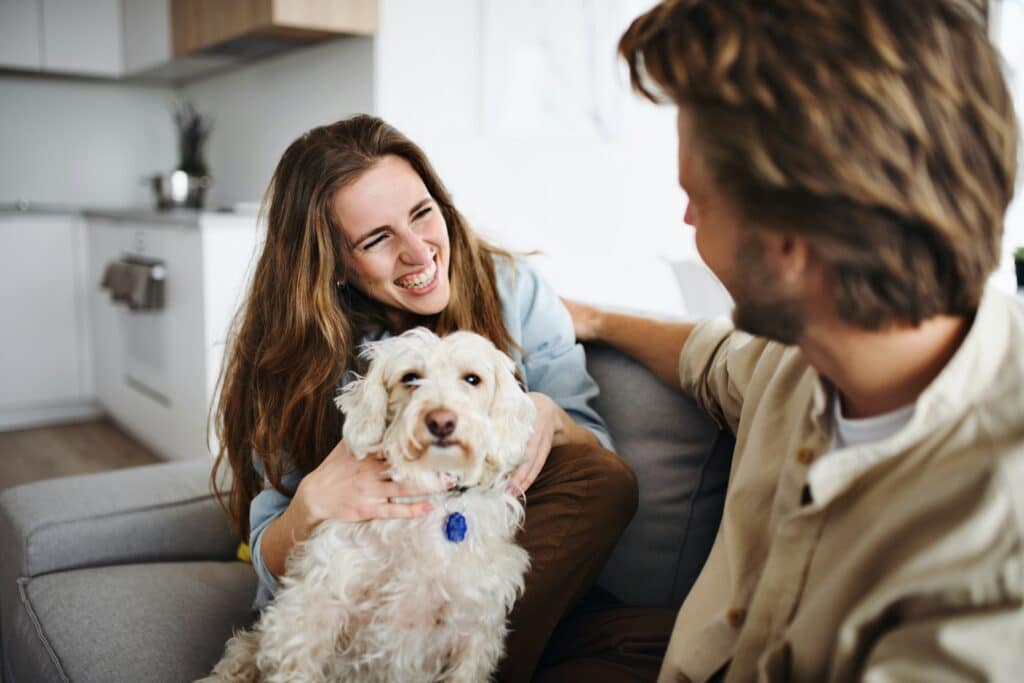 Junges Paar sitzt mit einem weißen Hund auf dem Sofa und sind glücklich.