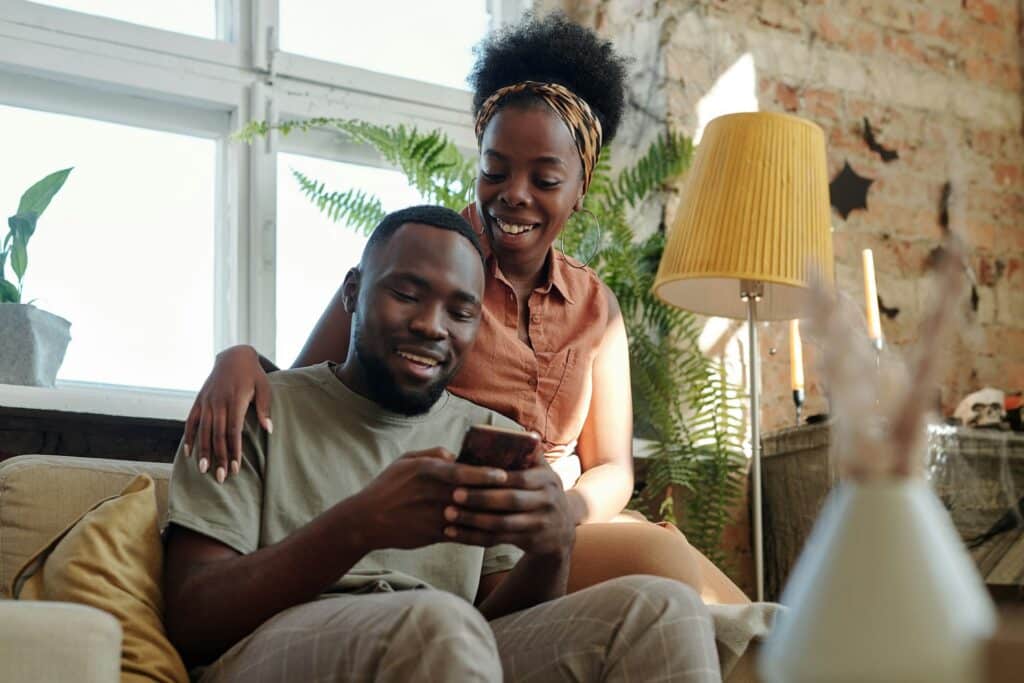 Fröhliches junges afrikanisches Paar in Freizeitkleidung schaut auf den Bildschirm des Smartphones, um nach einen Berater auf Berater Radar zu suchen