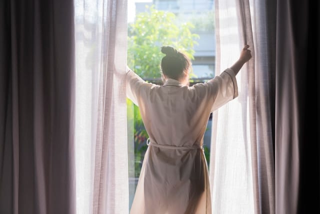 Frau öffnet morgens das Fenster in einem gepflegten Schlafzimmer.